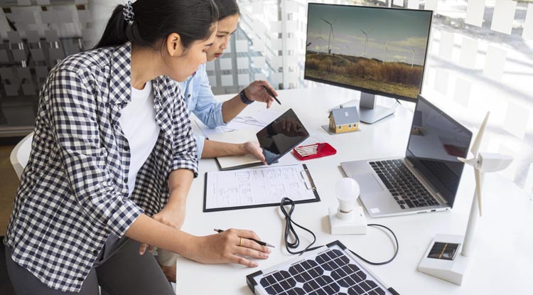 Duas profissionais de empresa analisando informações comerciais em tablet com outros dispositivos na mesa como notebook e monitor.