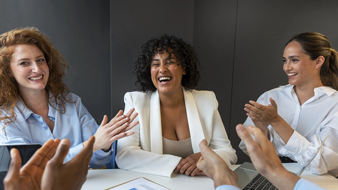 mulheres aplaudindo uma mulher por sua produtividade na empresa e seu sucesso com isso. A mulher do centro esta de branco e sorrindo, as duas mulheres dos dois cantas estão de azul e branco.