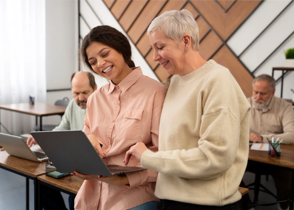 Duas mulheres, uma jovem e outra idosa, sorriem enquanto olham juntas para a tela de um notebook em um ambiente de sala de aula moderna. Atrás delas, dois homens mais velhos também estão concentrados em seus computadores. A cena transmite colaboração intergeracional e aprendizado em grupo.