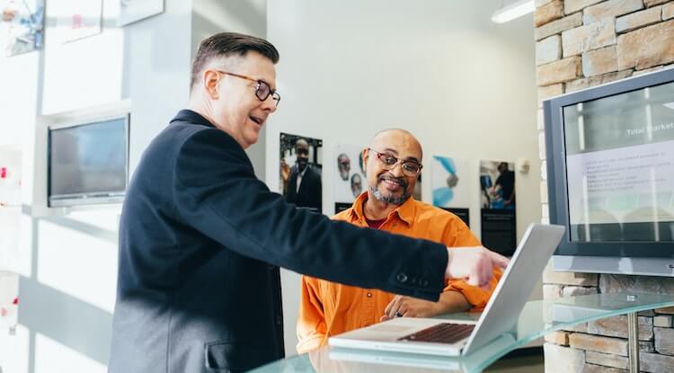 Duas pessoas olhando a tela de um computador, a pessoa da esquerda esta apontando para a tela do computador e está usando óculos de astes laranja, e vestindo um terno preto. a pessoa da direita está usando uma camisa laranja e óculos vermelho.