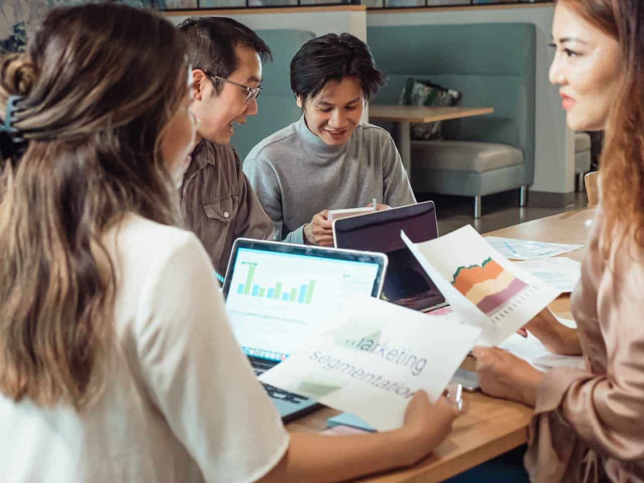 dois homens e duas mulheres trabalhando em clima amigável em torno de uma mesa com um notebook aberto e folhas com dados, todos eles são asiáticos.