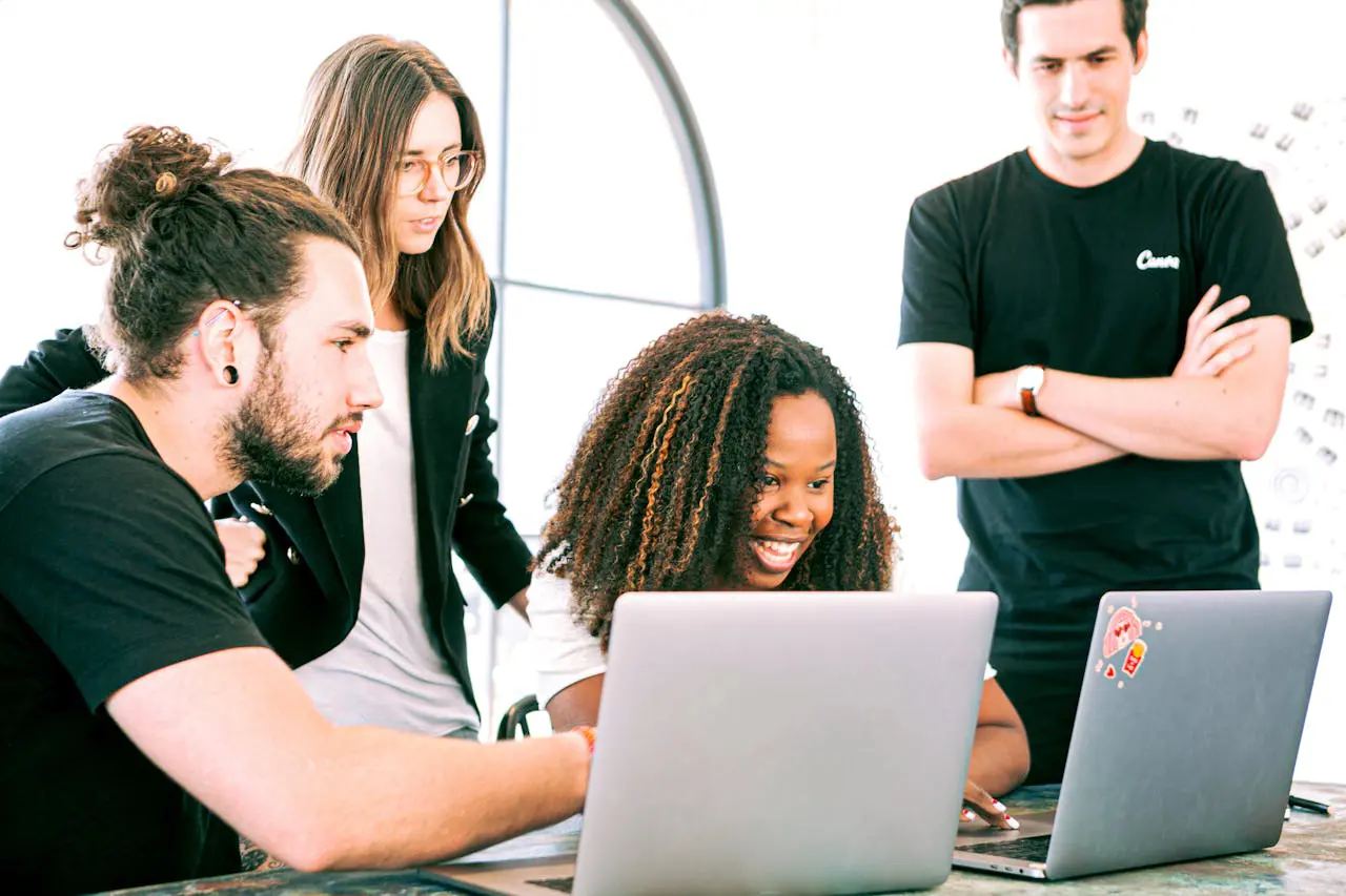 Um grupo de quatro jovens profissionais trabalha em equipe em um ambiente moderno e bem iluminado. Eles estão reunidos ao redor de dois notebooks, com expressões de entusiasmo e colaboração. Uma mulher de cabelos cacheados sorri enquanto digita no computador, enquanto os outros observam atentamente e interagem com o conteúdo na tela. O ambiente transmite um clima descontraído e colaborativo.