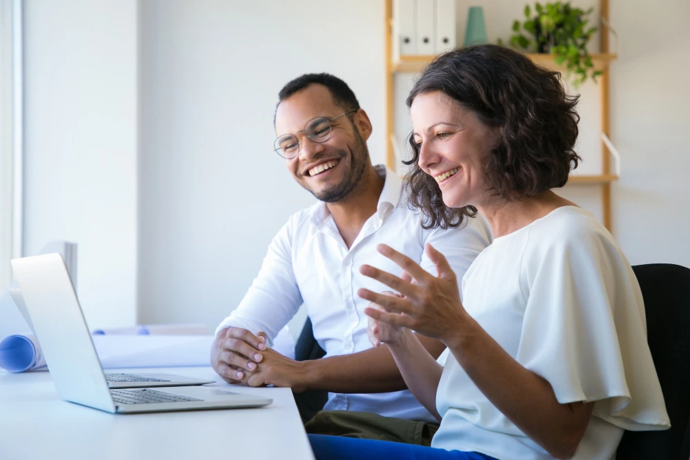 Um homem e uma mulher estão sentados em um escritório bem iluminado, interagindo de forma animada enquanto olham para a tela de um notebook. A mulher, de cabelos cacheados e blusa branca, gesticula com as mãos enquanto sorri, e o homem, de óculos e camisa branca, também sorri de maneira descontraída. O ambiente tem prateleiras ao fundo com plantas e itens de decoração, transmitindo uma atmosfera moderna e colaborativa.