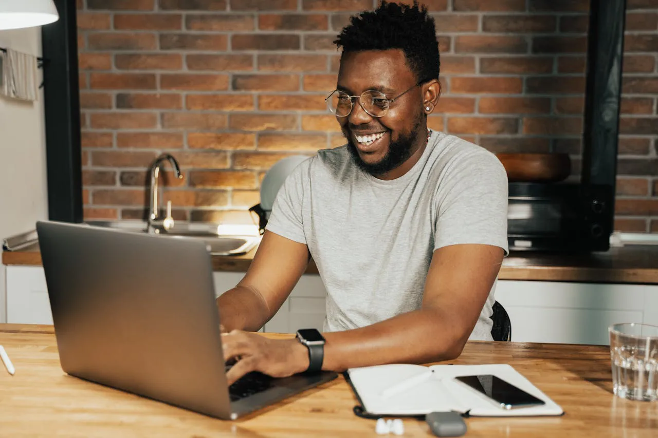 Um homem sorridente, de óculos e barba, está sentado em uma cozinha moderna com parede de tijolos aparentes, digitando em um laptop sobre uma mesa de madeira. Ele veste uma camiseta cinza e um smartwatch no pulso esquerdo. Ao lado do laptop, há um caderno aberto, um smartphone, um copo de água e fones de ouvido sem fio. O ambiente está bem iluminado, transmitindo uma atmosfera aconchegante e produtiva. Fazendo curso de Gamficação no setor educacional