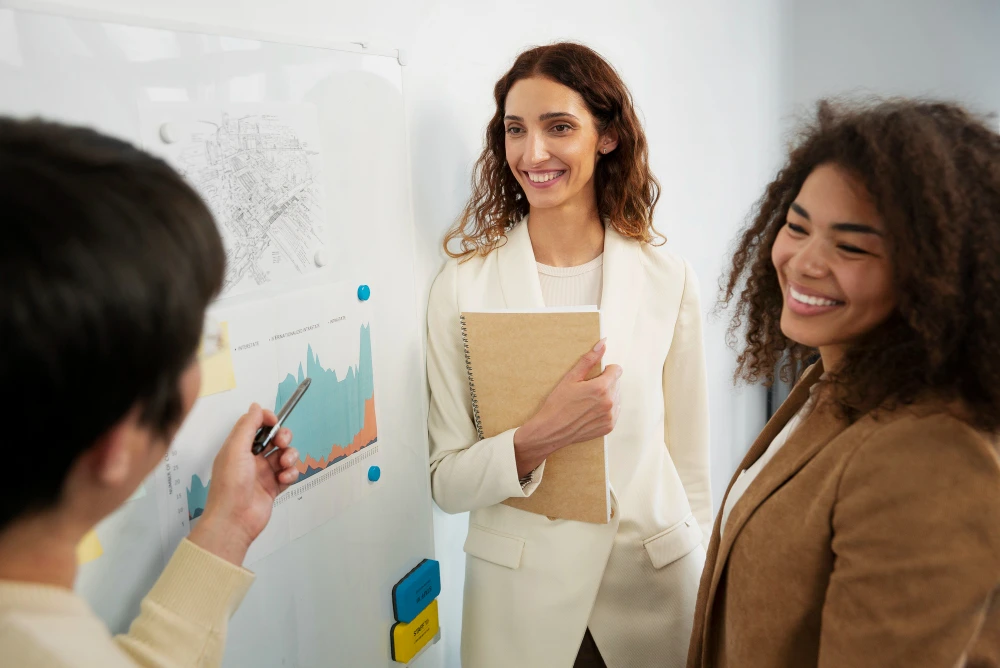 Três profissionais estão reunidos em um ambiente corporativo, discutindo dados apresentados em um quadro branco. Um homem, de costas para a câmera, aponta para um gráfico com uma caneta. Duas mulheres, vestindo blazers claros, sorriem e demonstram interesse na discussão. Uma delas segura um caderno. O ambiente sugere uma atmosfera colaborativa e produtiva.