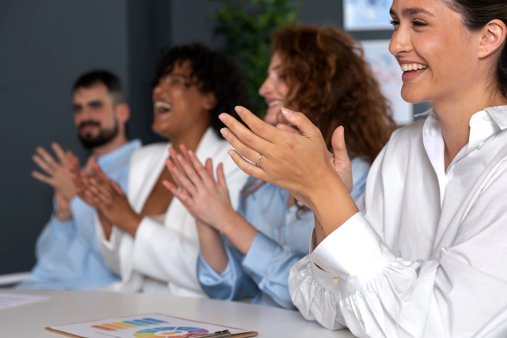 uatro pessoas sentadas lado a lado em uma mesa de reunião, sorrindo e aplaudindo com entusiasmo, demonstrando aprovação e espírito de colaboração em um ambiente profissional.