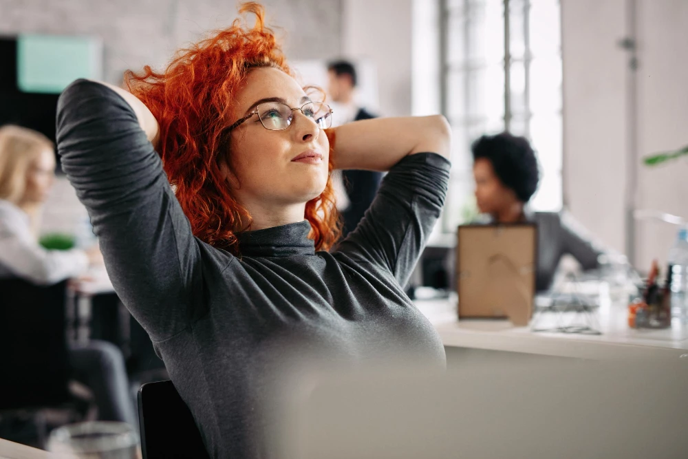 Mulher ruiva de óculos, sentada em um ambiente de escritório, com as mãos atrás da cabeça em uma postura relaxada e expressão de satisfação. Ao fundo, outras pessoas trabalham em suas mesas, em um ambiente colaborativo e iluminado.