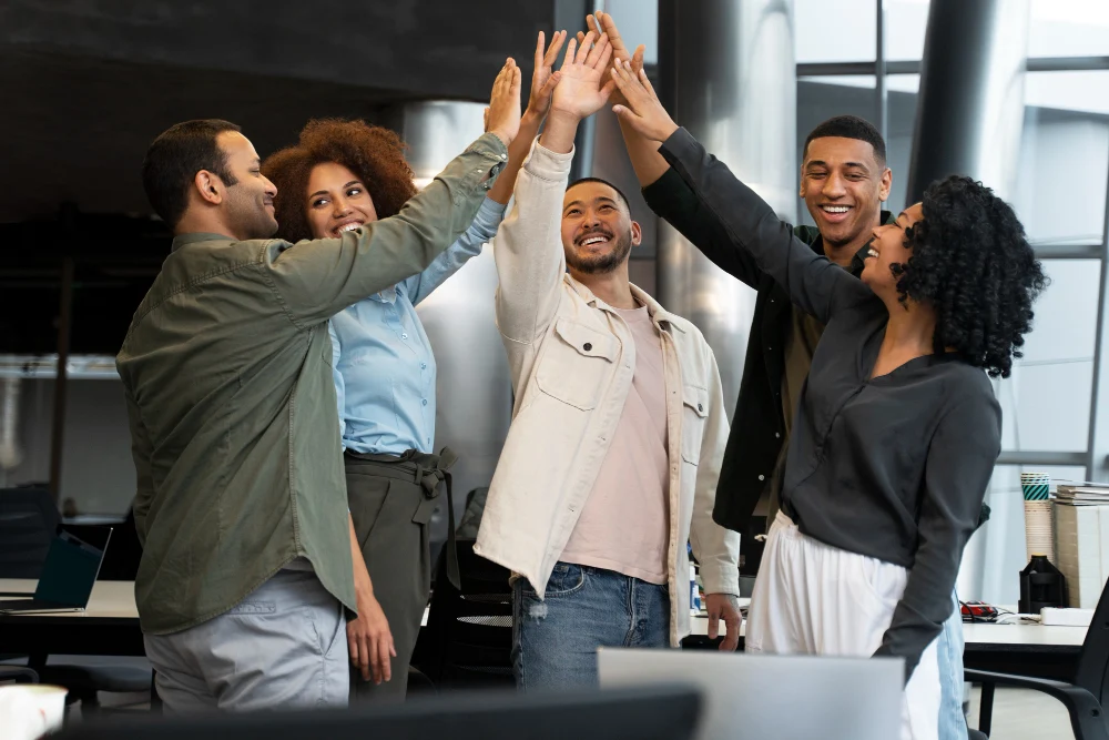 Grupo diverso de seis pessoas em um ambiente corporativo moderno realizando um high five coletivo, demonstrando espírito de equipe, celebração e colaboração. Todos estão sorrindo e visivelmente engajados, reforçando um clima positivo e motivador no local de trabalho. Ao fundo, vê-se parte de um escritório com mesas, cadeiras e equipamentos.
