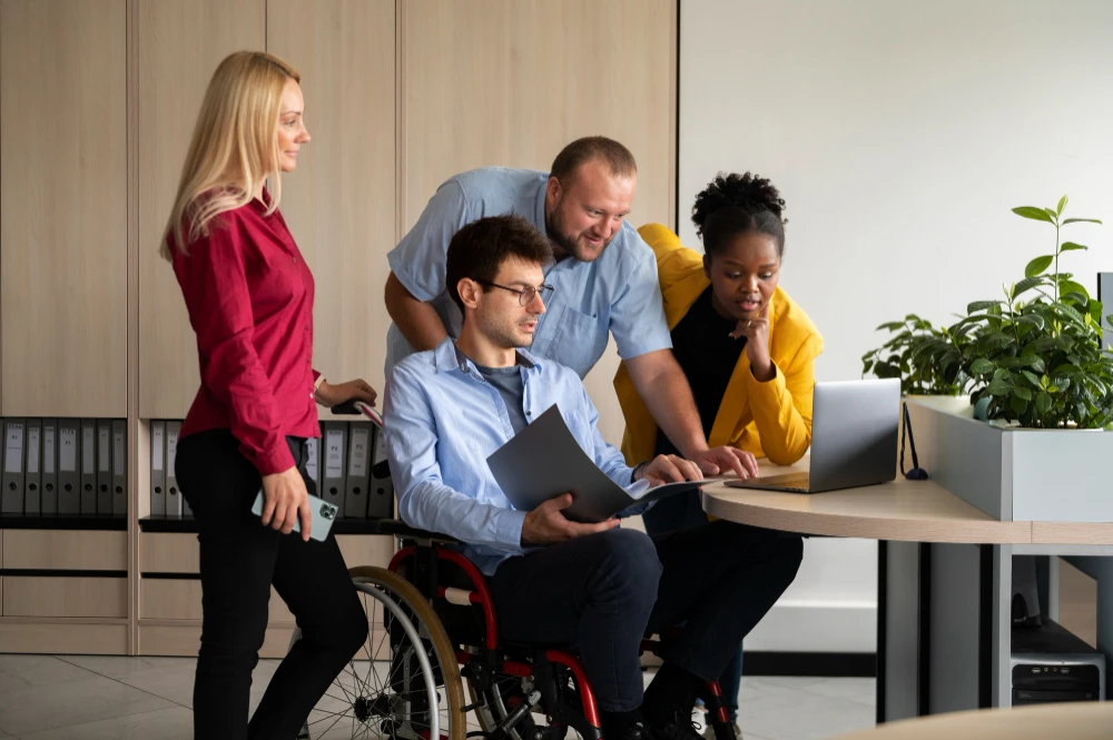 Quatro profissionais em um ambiente corporativo colaborando ao redor de uma mesa. Um homem em cadeira de rodas segura um laptop enquanto conversa com os colegas, que observam atentamente a tela. Uma mulher loira com blusa vermelha, um homem de camisa azul e uma mulher negra com blazer amarelo participam da reunião. Ao fundo, há um armário com pastas e uma planta sobre a mesa.