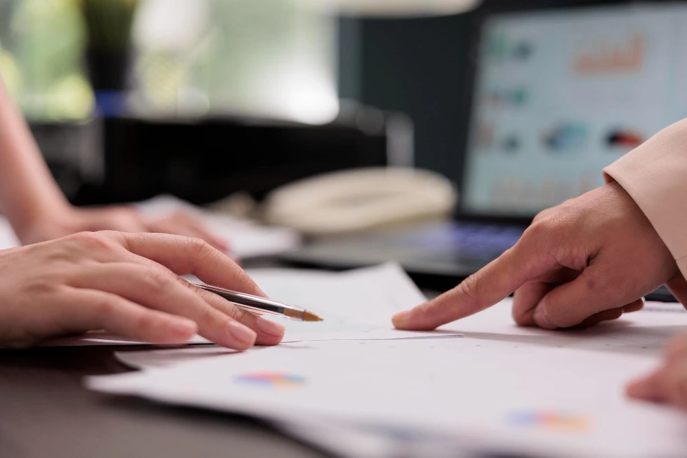 Duas pessoas analisam documentos impressos com gráficos coloridos sobre uma mesa. Uma delas segura uma caneta, enquanto a outra aponta para um dos papéis, sugerindo discussão ou tomada de decisão. Ao fundo, há um notebook com gráficos na tela, indicando um ambiente corporativo ou reunião de trabalho.