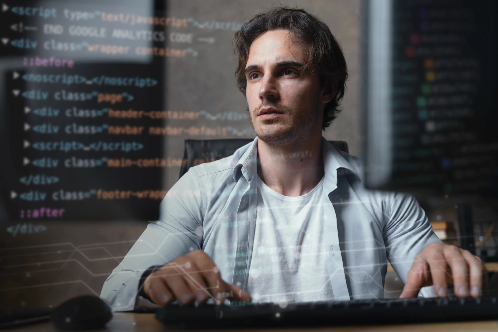 Homem jovem com expressão concentrada está sentado diante de dois monitores, digitando no teclado e utilizando um mouse. Ele usa uma camiseta branca e camisa azul clara aberta, e está cercado por códigos de programação sobrepostos na imagem, indicando que está desenvolvendo ou analisando software. O ambiente é escuro e moderno, com foco total em seu trabalho na área de tecnologia.
